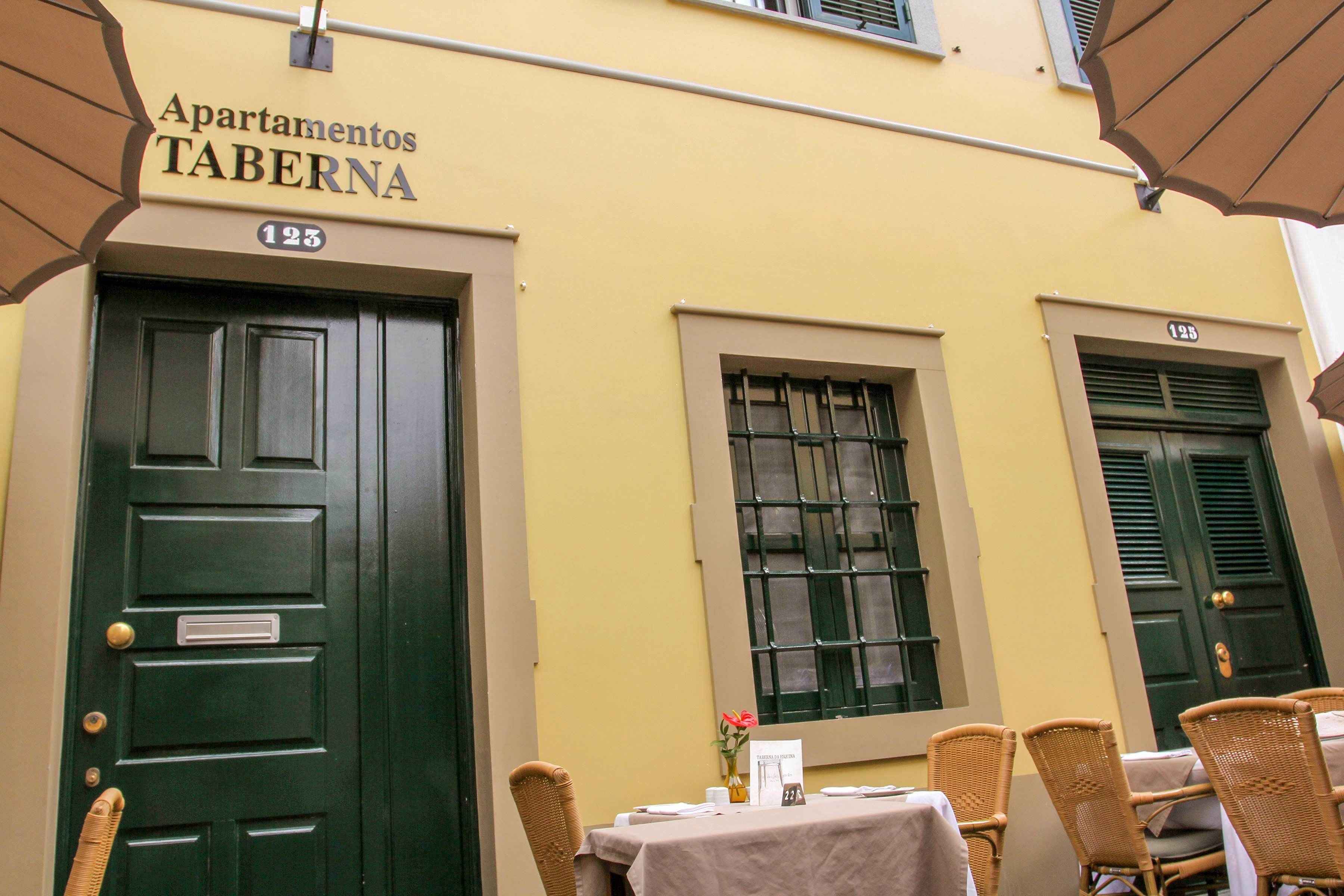 A building with the sign 'Apartamentos TABERNA' and the number '123'. There are outdoor tables and chairs, a small vase with flowers, and a menu sign.
