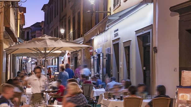 A bustling outdoor dining area with tables and chairs, surrounded by buildings with shops and signs. - Apartamentos Taberna (Funchal)