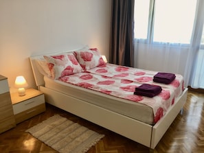 Cama bem arrumada com colcha florida, abajur e mesinha de cabeceira com luminária.