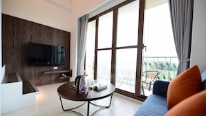 A modern living room with a flat-screen TV mounted on a wooden wall, a round coffee table with books and a mug, a sofa with cushions, and large windows with curtains. - Yasengawa (Hengchun)