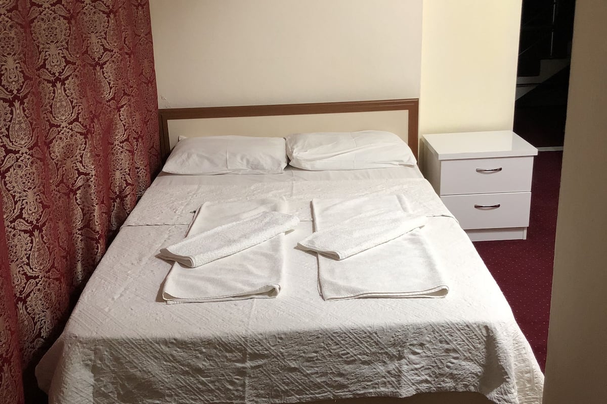 A neatly made bed with white linens and towels folded on top, a white bedside table, and a red patterned curtain.