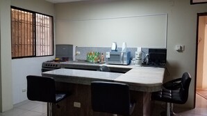 A compact kitchen with a central island, two bar stools, a gas stove, a microwave, a coffee maker, and a window with bars. - Casa Serena (Guayaquil)