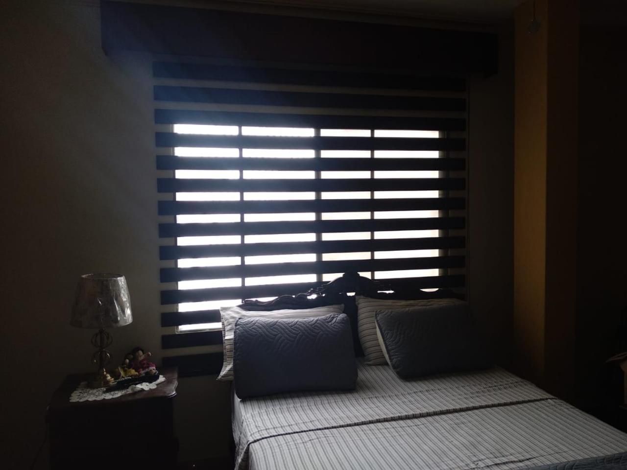 Un dormitorio con cama, mesita de noche, lámpara y ventana con persianas.