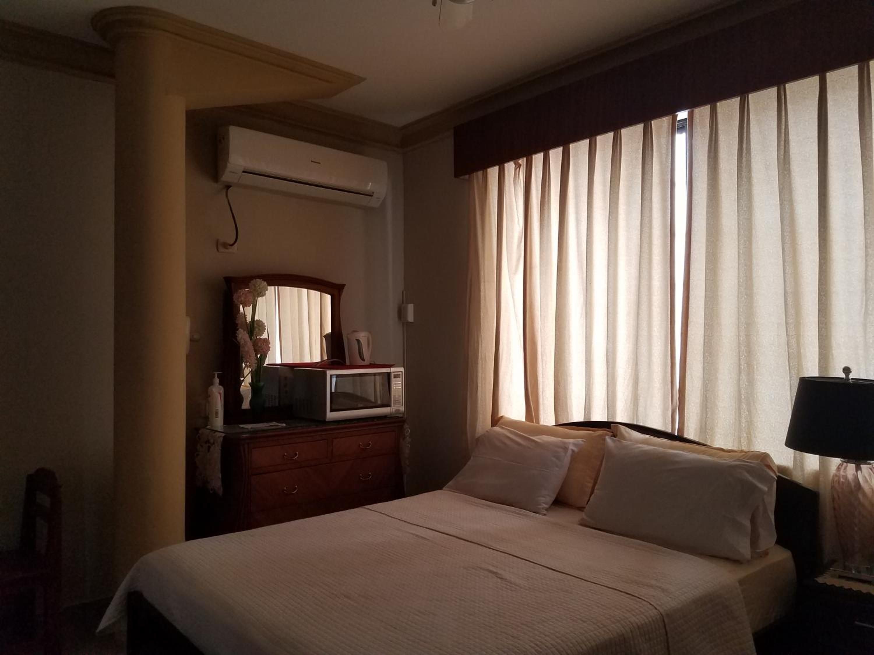 Una habitación de hotel con cama, cómoda, aire acondicionado, ventana con cortinas y una lámpara.