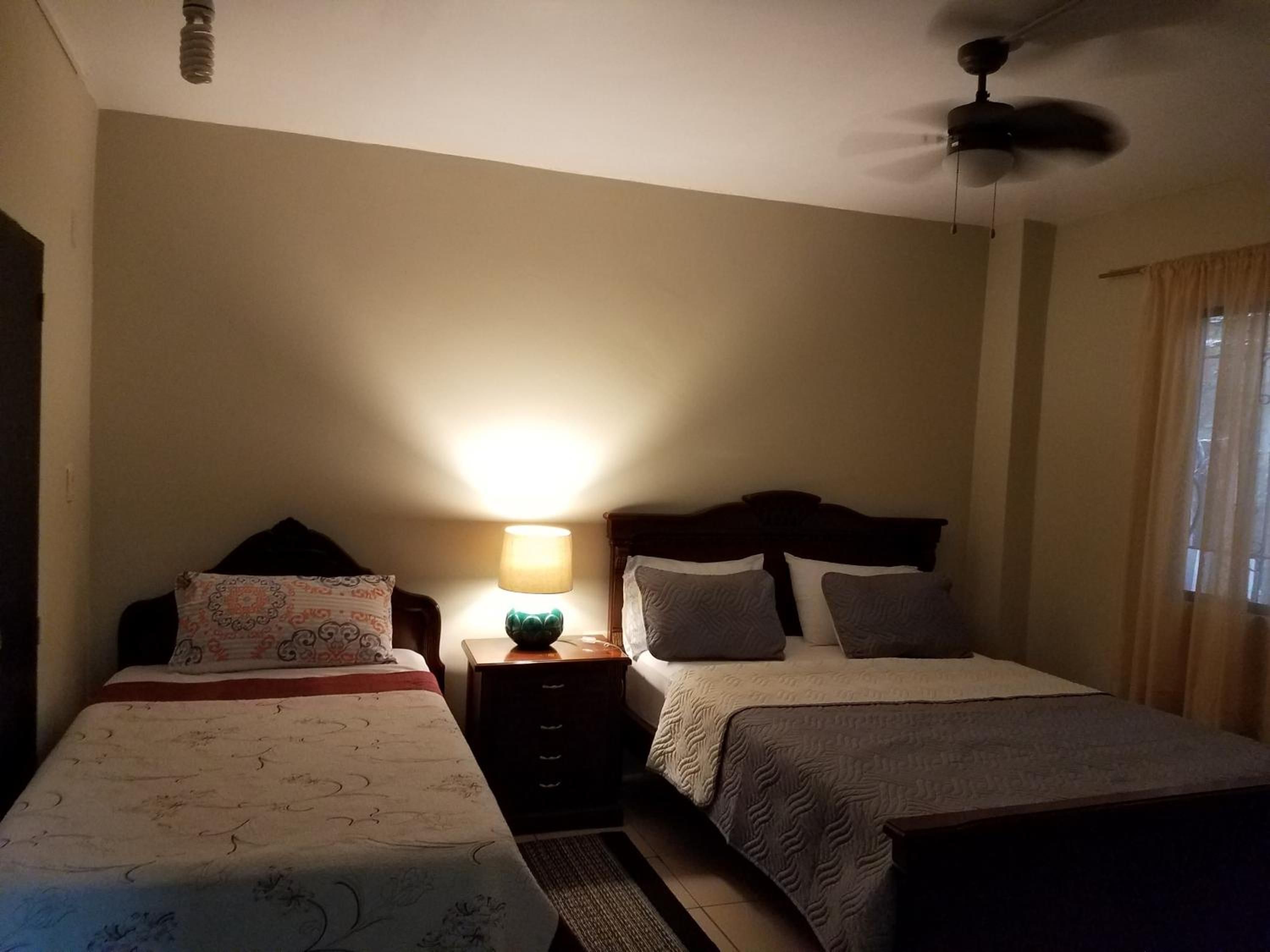 Camera d'albergo con due letti, un comodino con lampada, una ventola a soffitto e una finestra con tende.