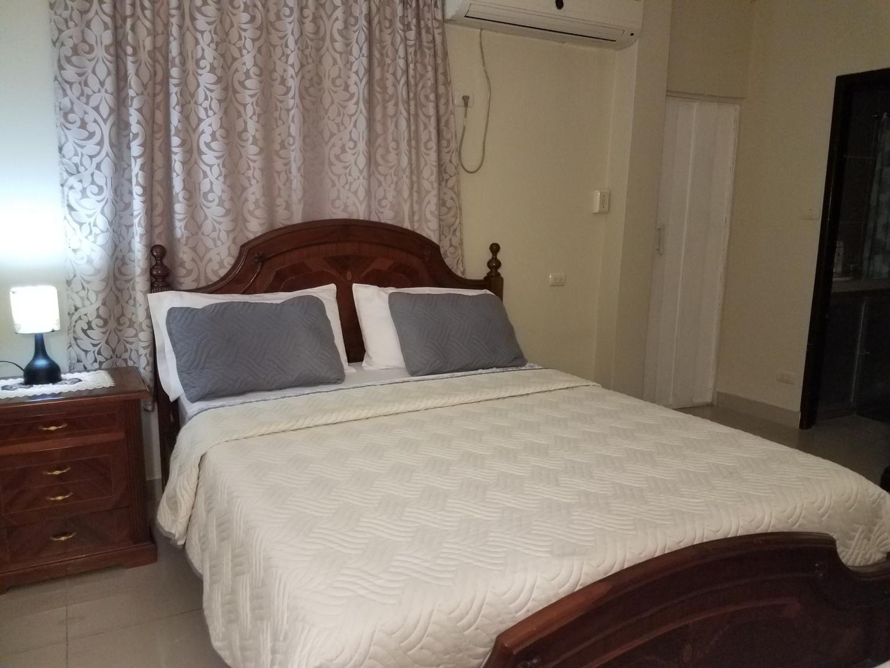 Un dormitorio con cama de madera, ropa de cama blanca, una mesita de noche con lámpara y cortinas estampadas.
