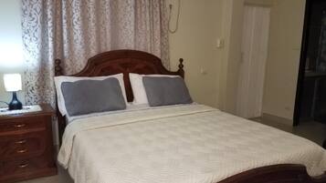 Un dormitorio con cama de madera, ropa de cama blanca, una mesita de noche con lámpara y cortinas estampadas.