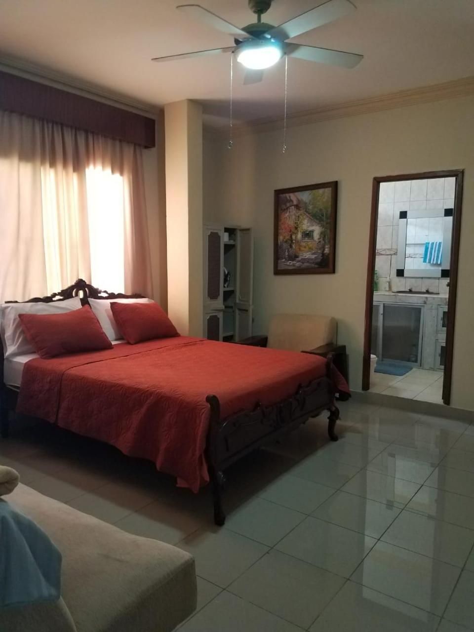 Un dormitorio con cama, ventilador de techo, un cuadro y vista a la cocina.
