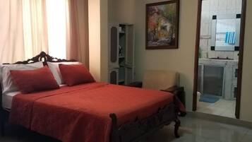Un dormitorio con cama, ventilador de techo, un cuadro y vista a la cocina.