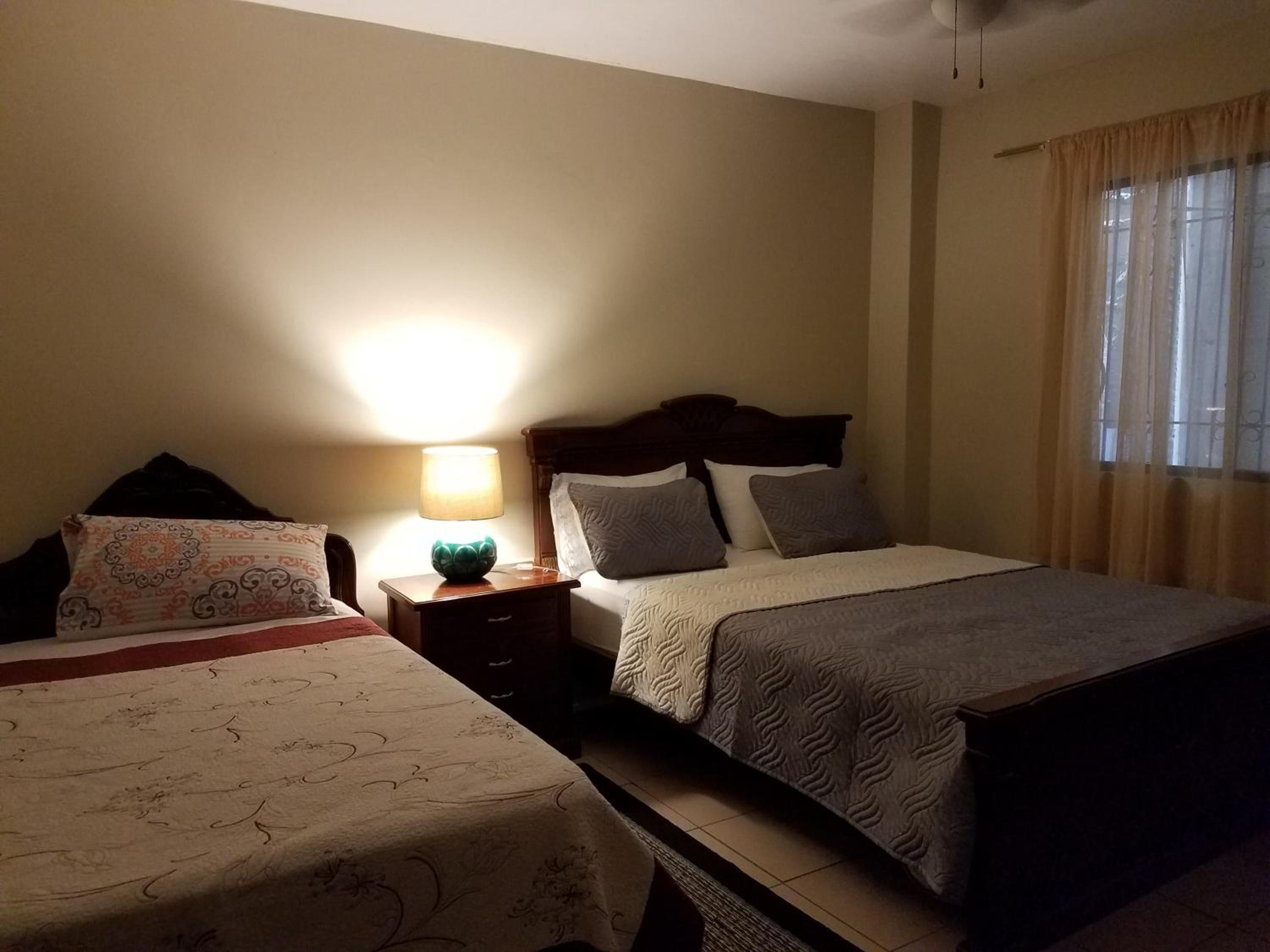 Quarto de hotel com duas camas, uma mesa de cabeceira com luminária e uma janela com cortinas.