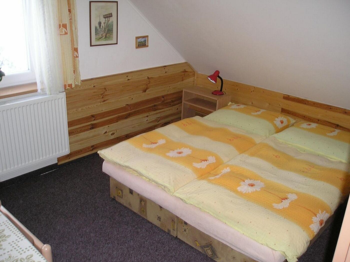 Une chambre à coucher avec une tête de lit en bois, un lit simple recouvert d’une couvre-lit à motifs floraux, une table de chevet avec une lampe et un radiateur.