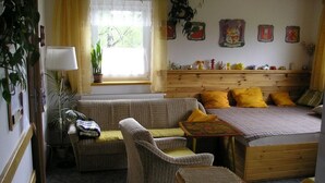 A cozy room with a bed, a sofa, a small table, and a bench. There is a window with curtains, a plant, and wall decorations.