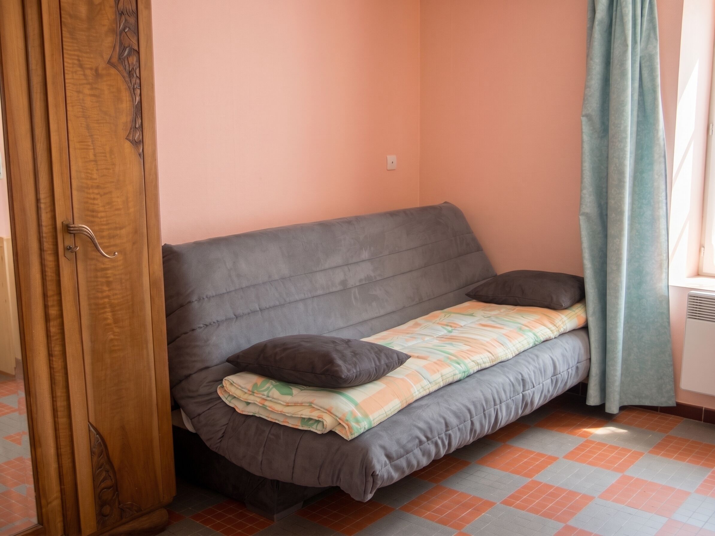 A small room with a grey sofa, a wooden door, a window with a curtain, and a tiled floor.