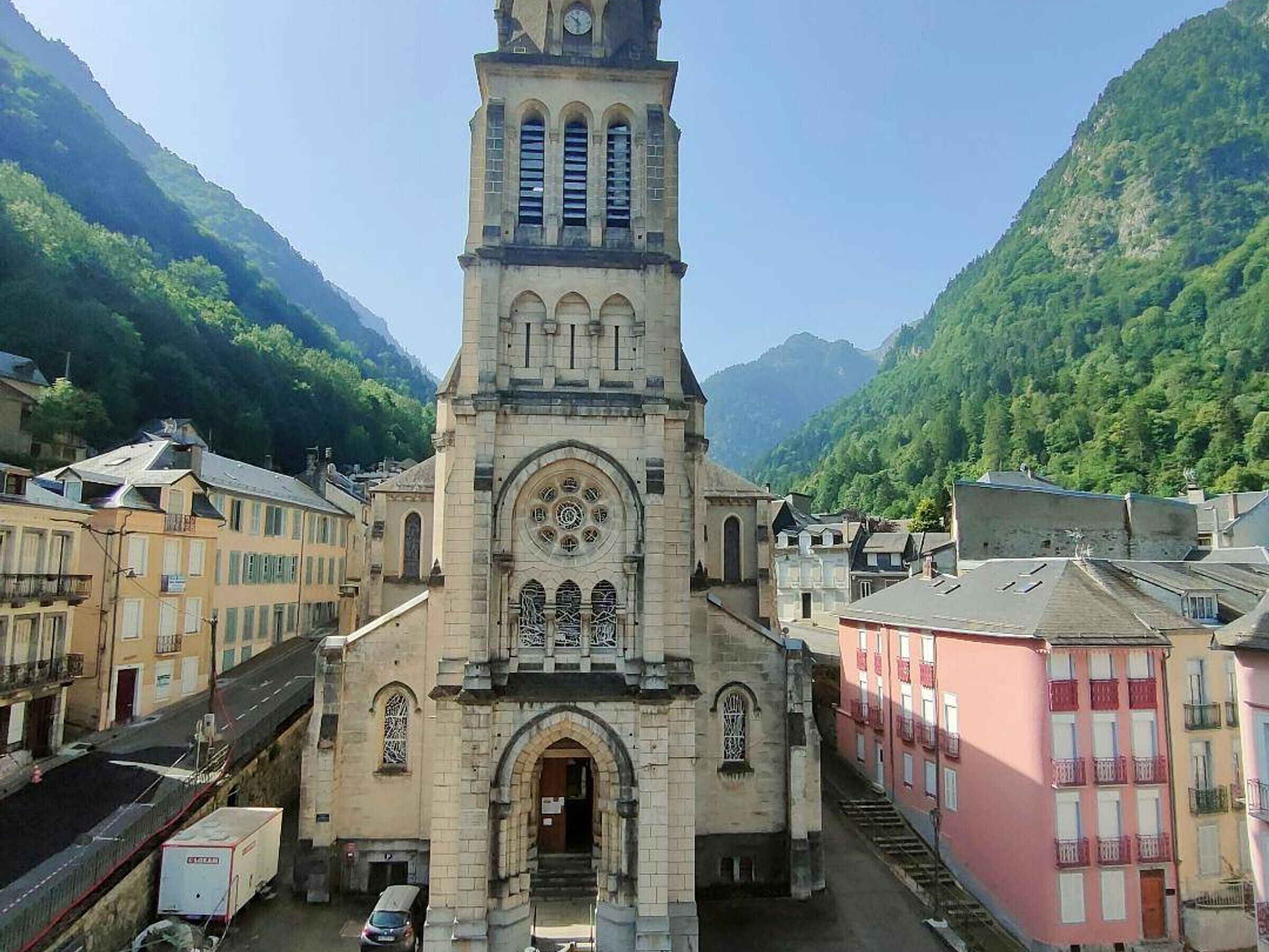 A historic church with a clock tower, surrounded by buildings and a mountainous backdrop.