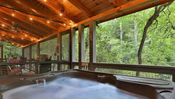 A hot tub with a wooden deck and large windows overlooking a forest. - River Rhapsody | Ellijay, GA (Ellijay)