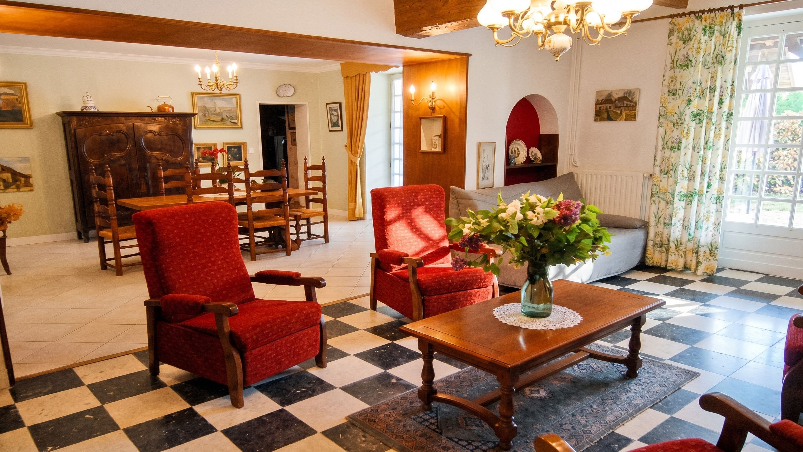 A room with a checkered floor, a wooden table with a vase of flowers, and a red armchair.