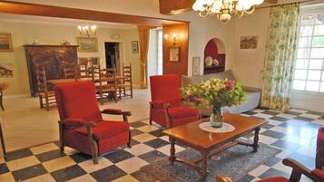 A checkered floor, two red armchairs, a wooden coffee table with a vase of flowers, and a dining area with chairs and a wooden cabinet.