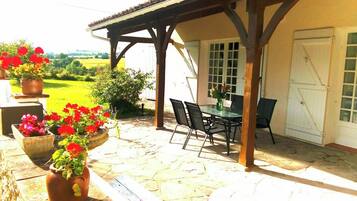 A covered patio with a stone floor, a table with chairs, and potted flowers.