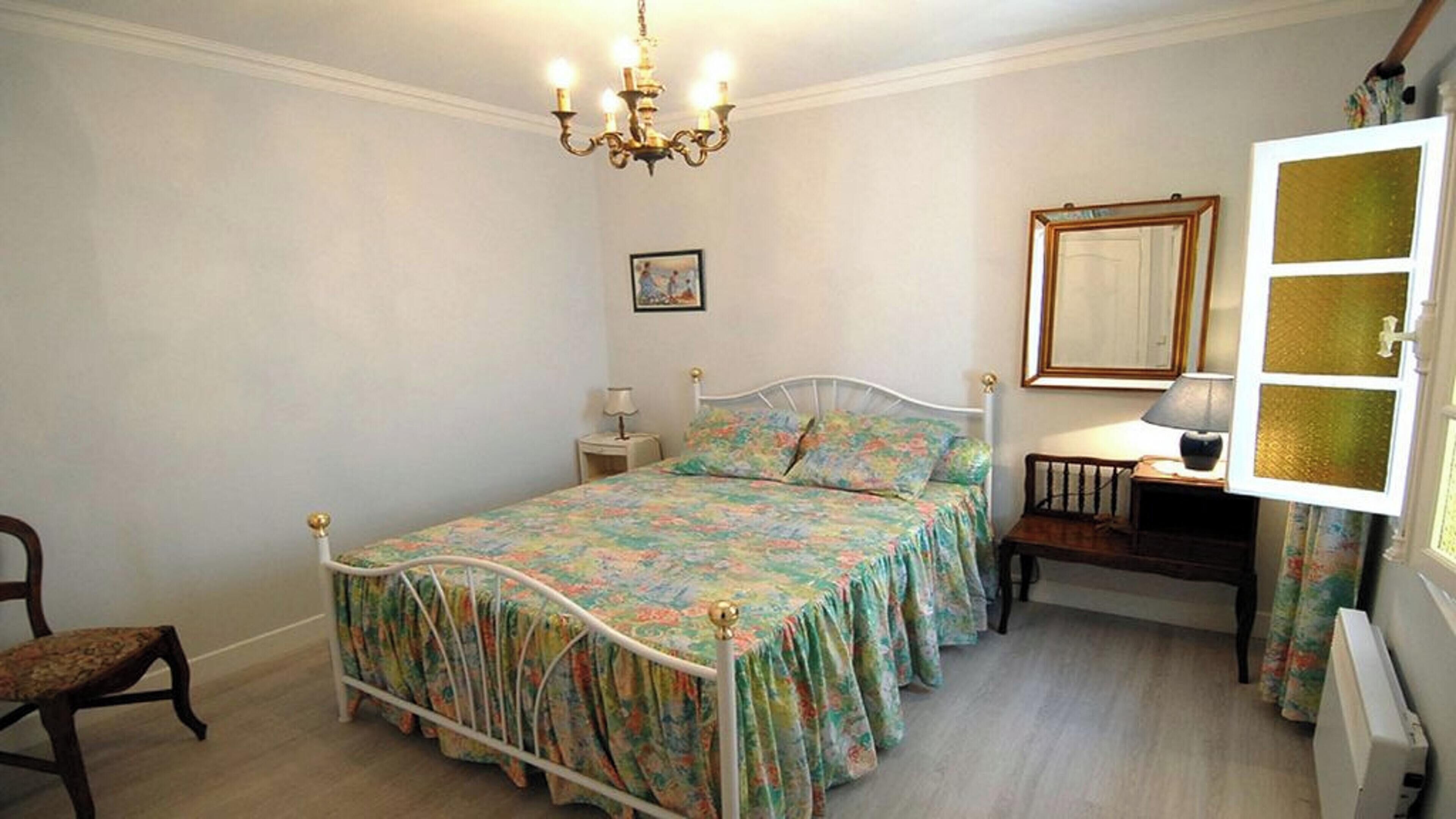 Een slaapkamer met een bed, nachtkastje, stoel, dressoir, spiegel en een kroonluchter.