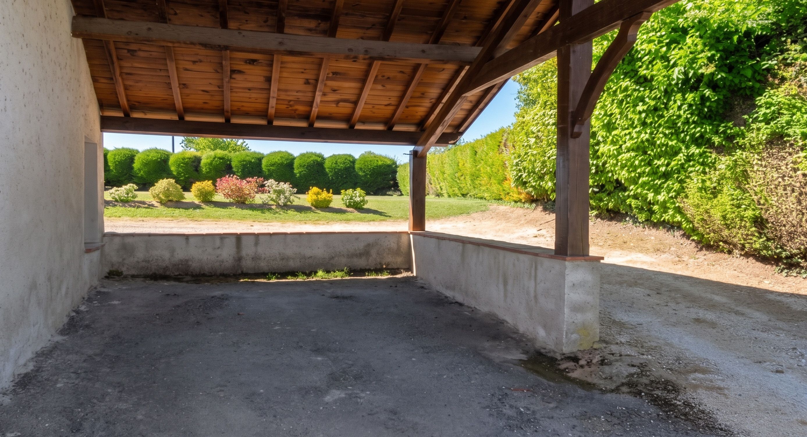 An empty room with a concrete floor, wooden ceiling beams, and a view of a landscaped garden with various shrubs and trees.