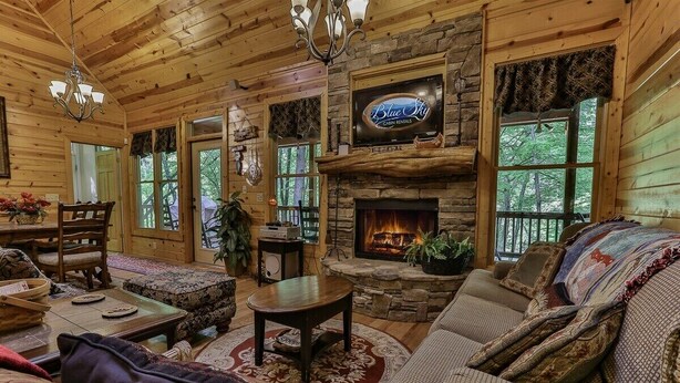 A log cabin interior with a stone fireplace, a television mounted above it, and a cozy seating area with a sofa and armchairs.