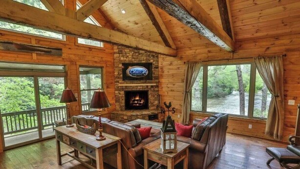 Ein Blockhaus-Interieur mit Kamin aus Stein, Holzmöbeln und großen Fenstern mit Blick auf einen Wald.