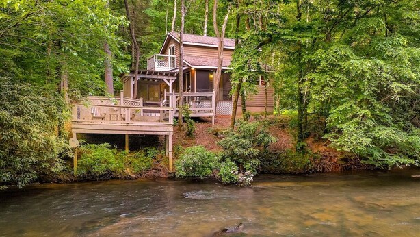 A cabin with a deck overlooking a river, surrounded by dense forest. - Rendezvous on the River | Ellijay, GA (Ellijay)