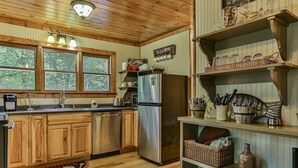 A kitchen with wooden cabinets, a stainless steel refrigerator, and a window with a view of trees. - Rendezvous on the River | Ellijay, GA (Ellijay)