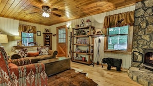 A rustic cabin interior with a wooden ceiling, a stone wall, a fireplace, a bookshelf, and a cowhide rug. - Rendezvous on the River | Ellijay, GA (Ellijay)