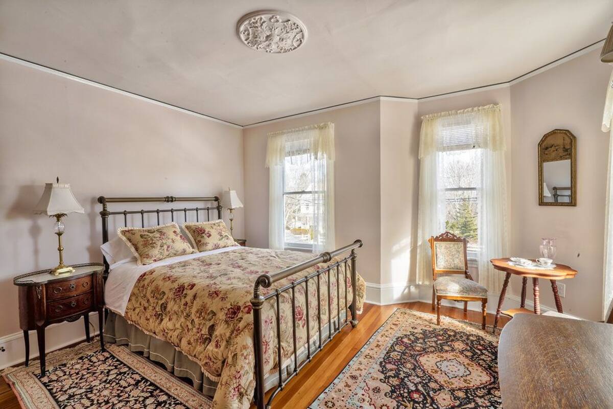 A bedroom with a four-poster bed, a nightstand with a lamp, two chairs, a small table, and a mirror on the wall.
