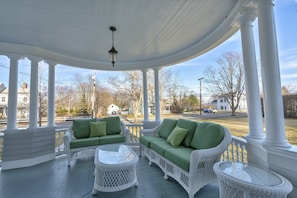 A covered porch with white columns, green cushioned seating, and a glass table. - Victorian Seasons Stirling (Greenport)