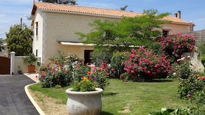 A two-story house with a tiled roof, surrounded by a well-maintained garden with various flowering plants and a large planter. - Aux Orangers (Luxe)