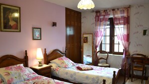 A bedroom with two wooden beds, a nightstand with a lamp, a wooden chair, and a window with curtains. - Aux Orangers (Luxe)