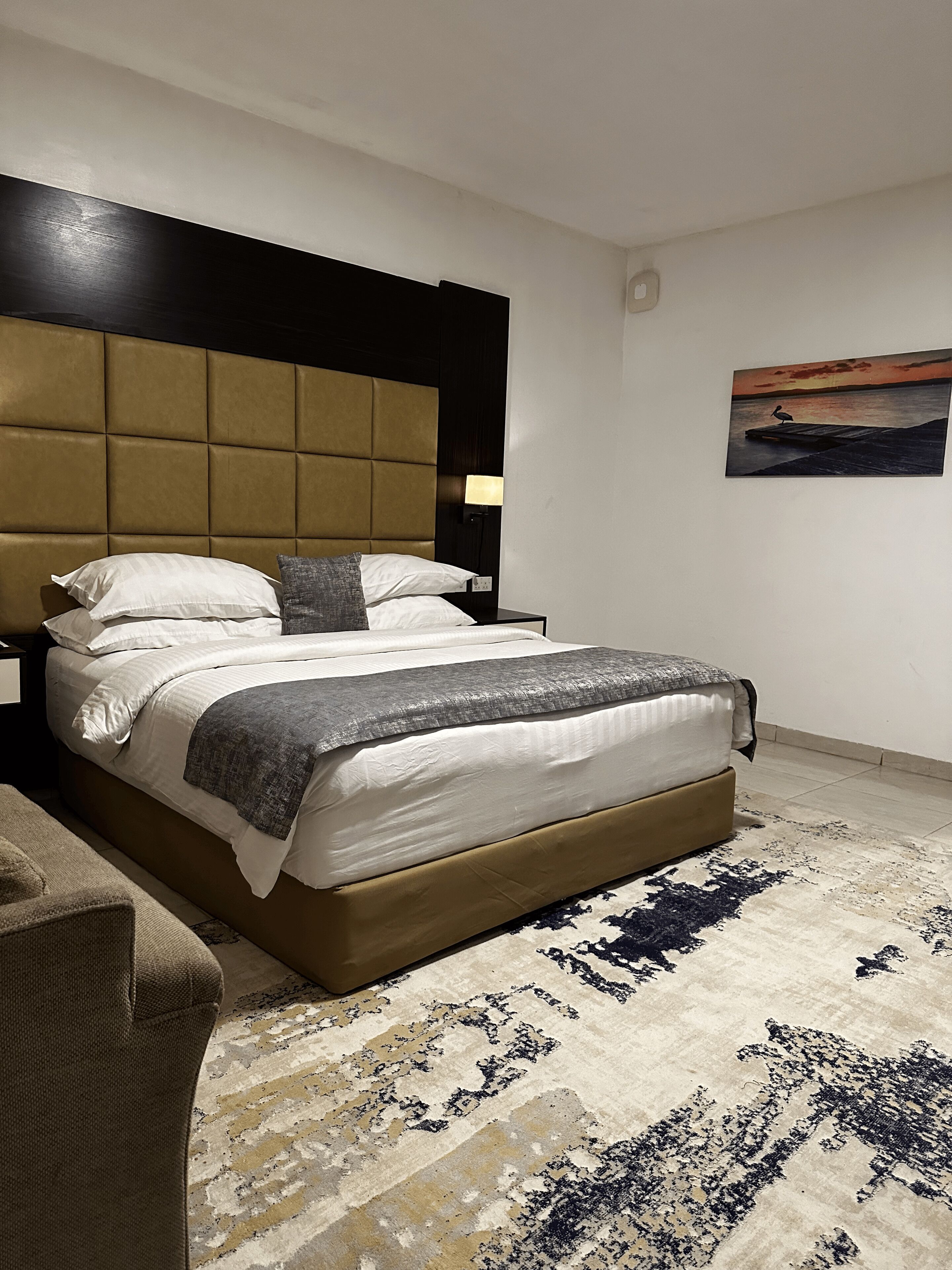 A modern hotel room with a large bed, a headboard, a nightstand, a lamp, and a painting on the wall.