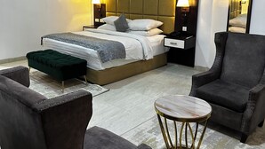 A modern hotel room with a large bed, two bedside tables, a nightstand, a green ottoman, two armchairs, and a small round table. - Rollace Hotel Lagos INTL (Lagos)