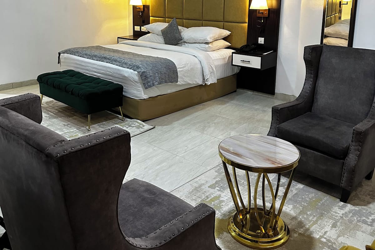 Una habitación de hotel moderna con una cama grande, dos mesitas de noche, un taburete verde, dos sillones y una pequeña mesa redonda.