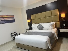 A hotel room with a large bed, a headboard with a light brown panel, two bedside tables with lamps, a nightstand, a wall-mounted air conditioner, and a painting of a sunset over water.