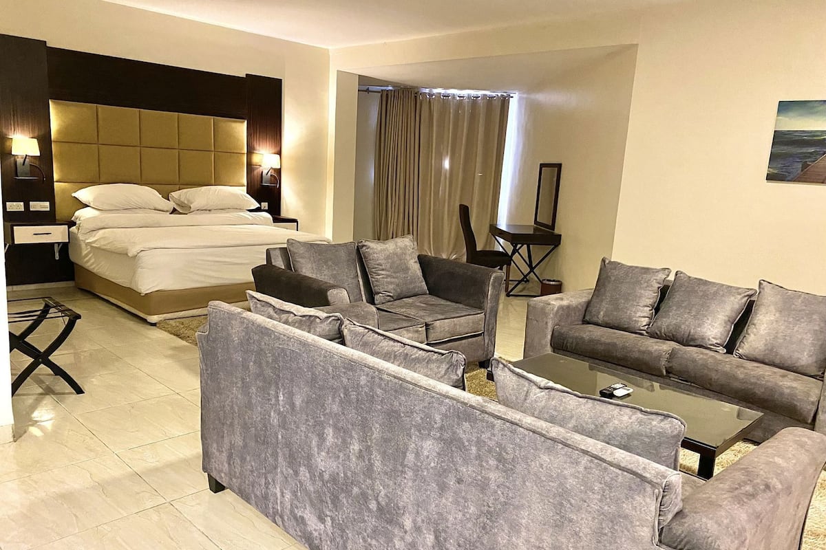 Habitación de hotel con cama, dos sofás, una mesita de centro, una mesita auxiliar y un amplio ventanal con cortinas.