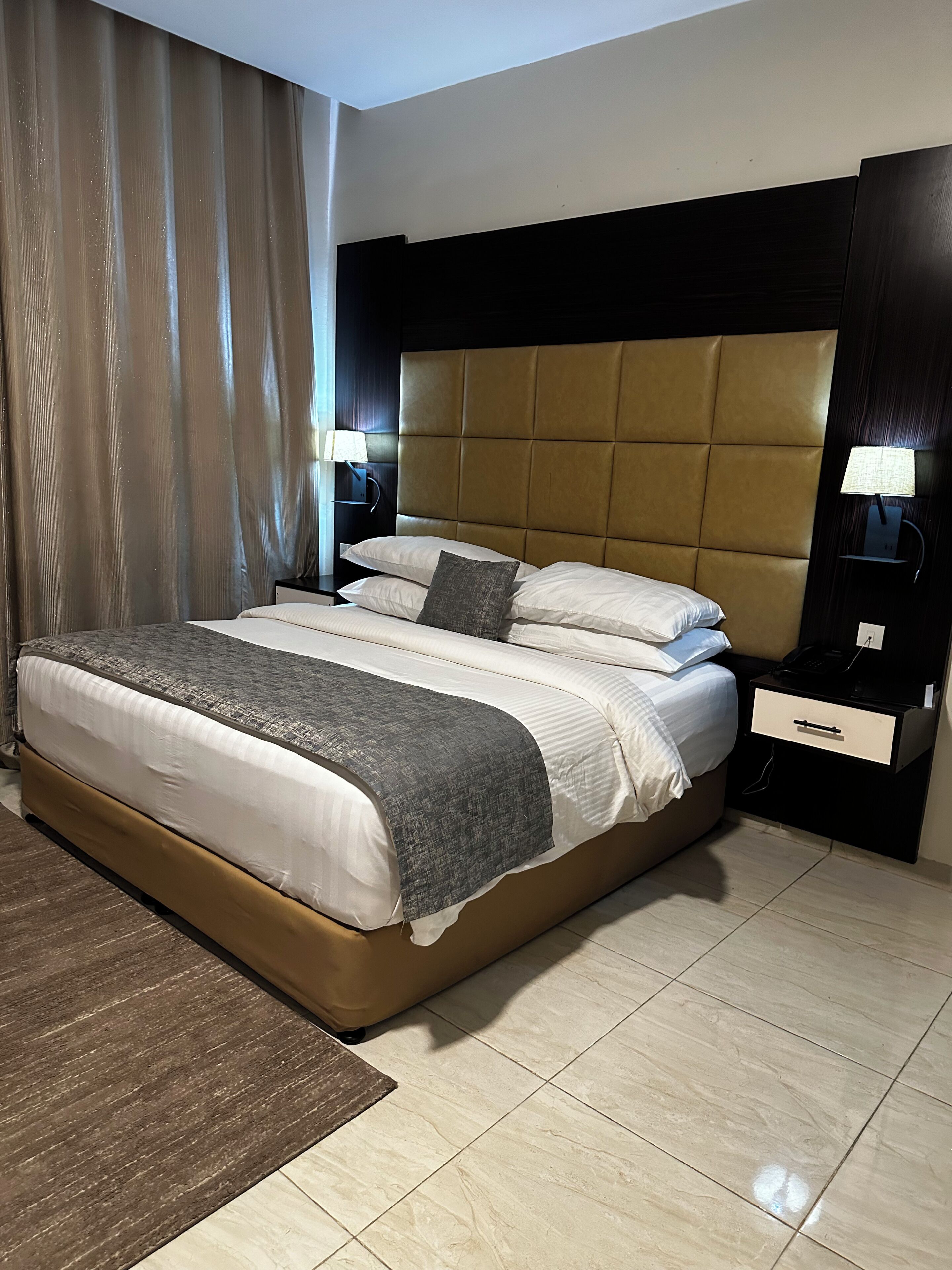 A hotel room with a large bed, two bedside tables, a nightstand, a lamp, and a window with curtains.