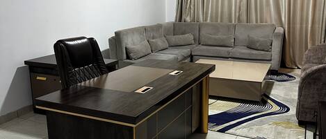 A modern living room with a dark wooden desk, a grey sofa, and a large window with beige curtains.