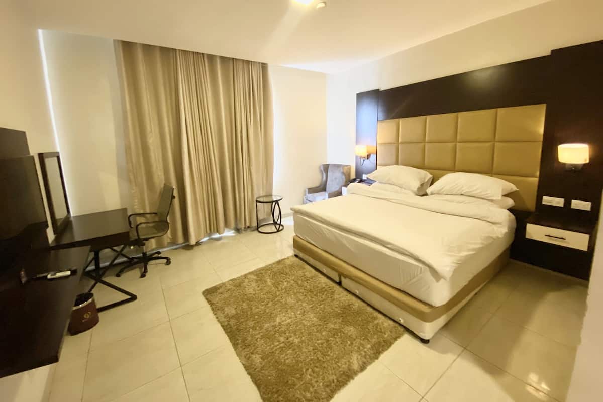 Habitación de hotel con cama, escritorio, silla, televisor, lámpara y ventana con cortinas.