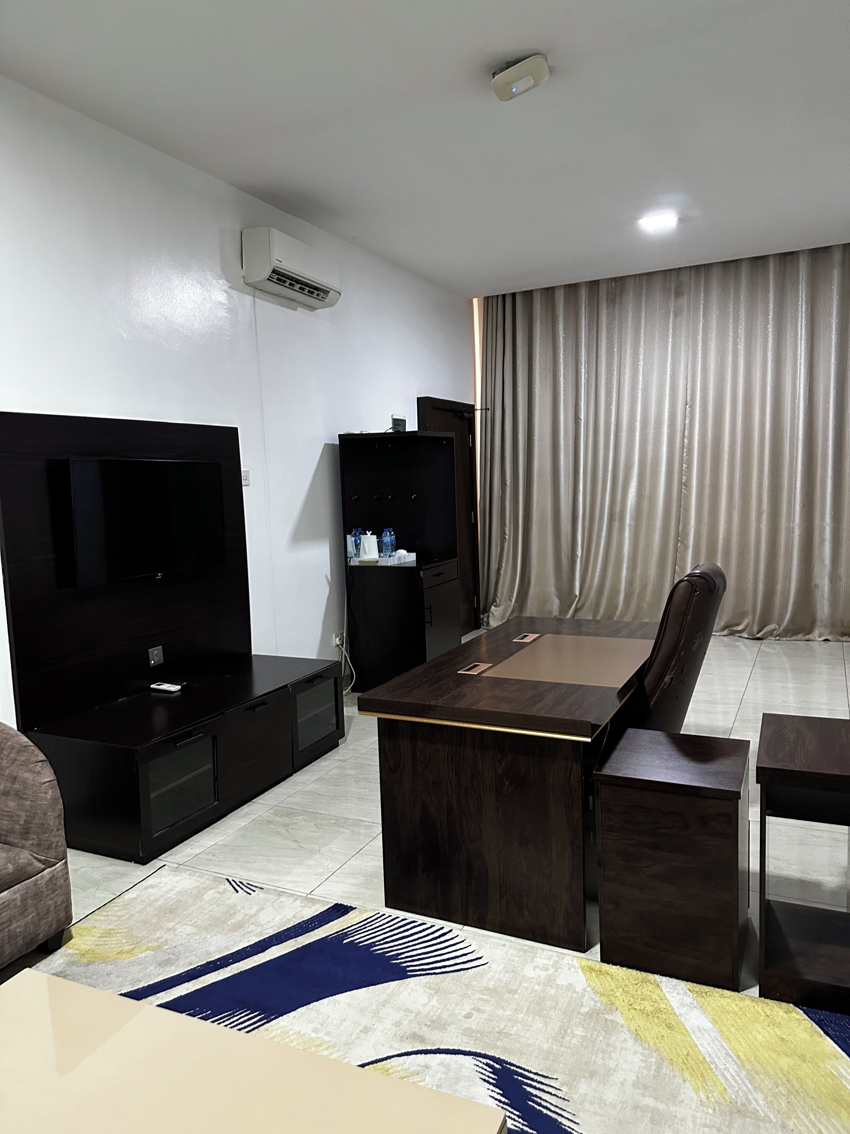 A modern living room with a flat-screen TV, a wooden coffee table, a sofa, a dining table with chairs, a black entertainment unit, and a large window with curtains.