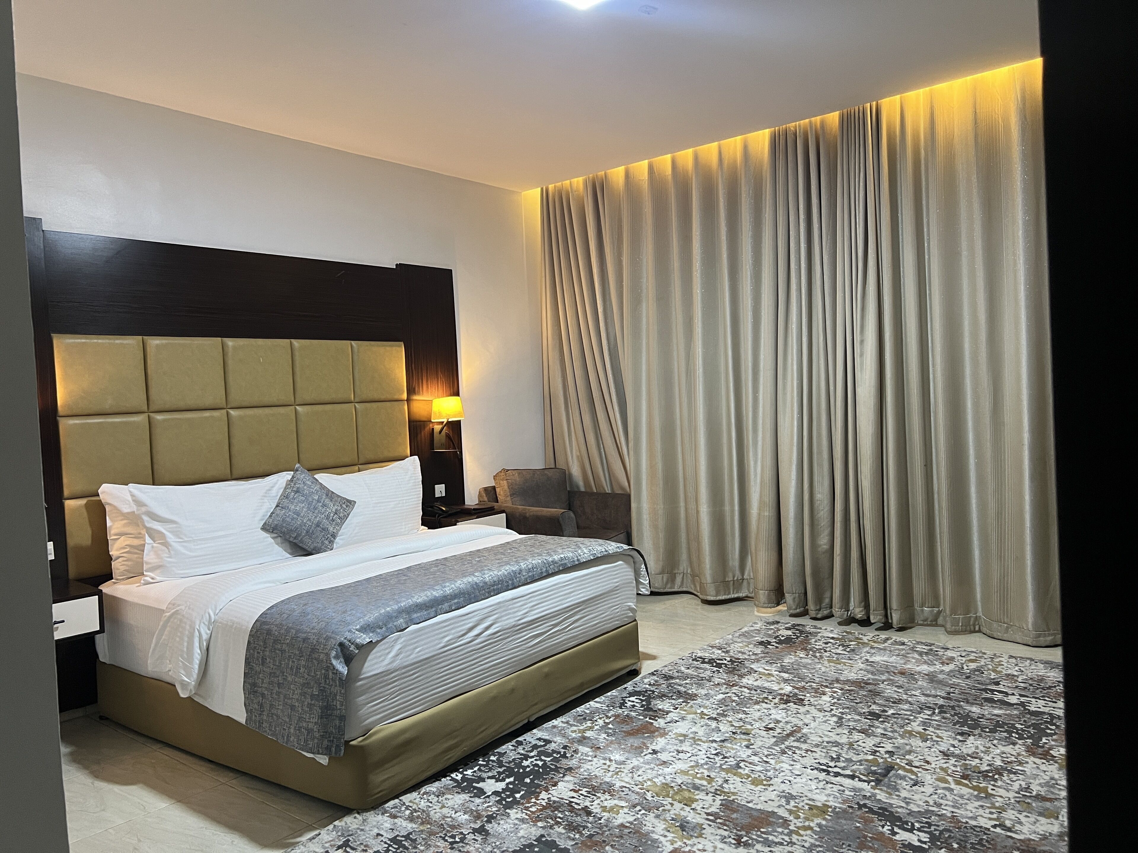 Quarto de hotel com uma cama grande, mesa de cabeceira, luminária, cadeira e vista para o exterior através de cortinas.