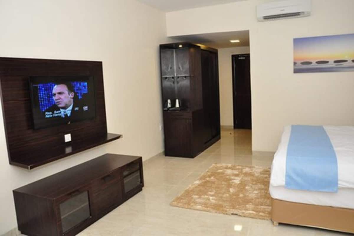 Una habitación de hotel con una televisión montada en la pared, una cama con manta azul, un armario de madera y una alfombra beige.