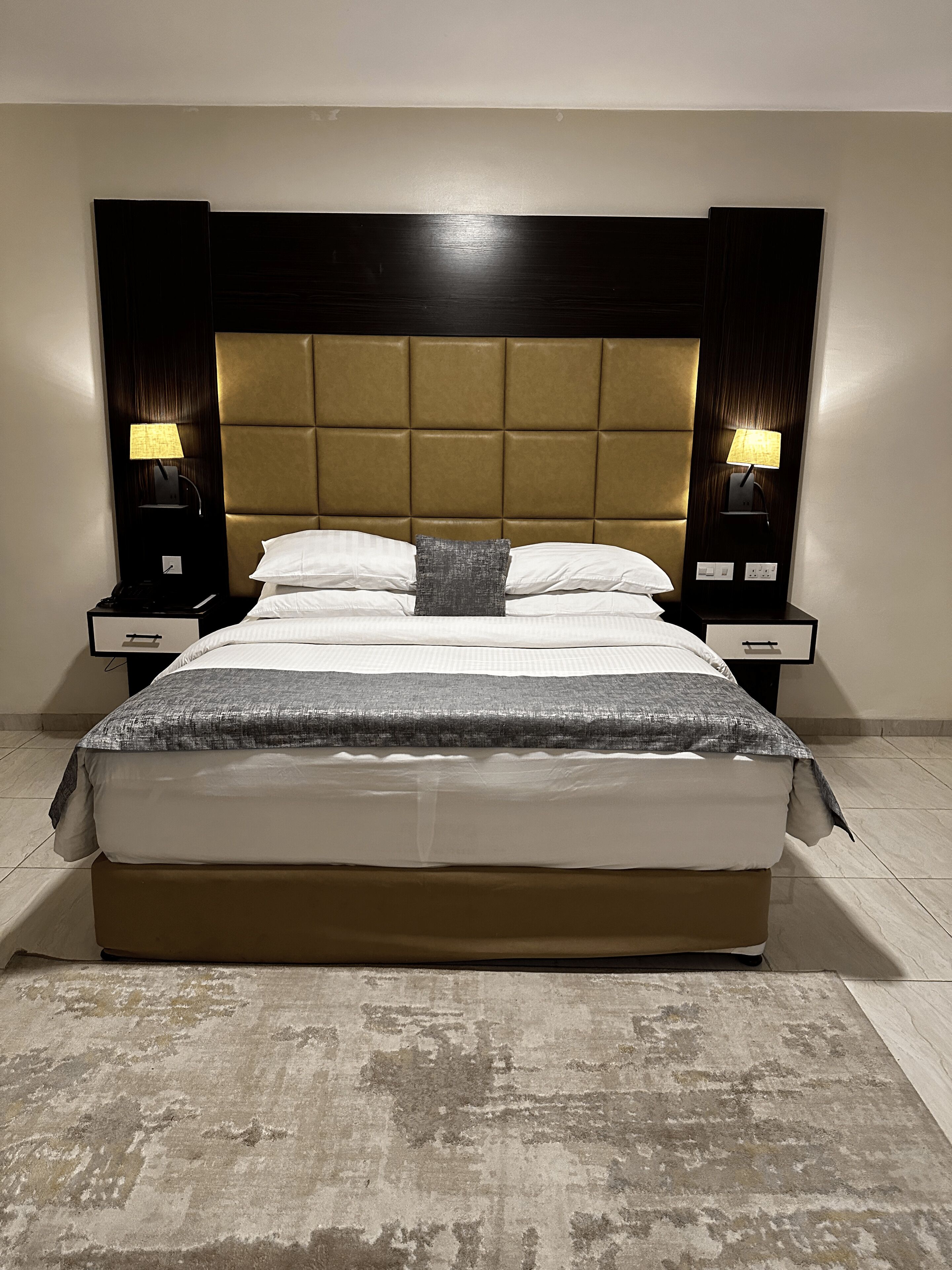 A large bed with a tufted headboard, two bedside tables with lamps, and a patterned rug.
