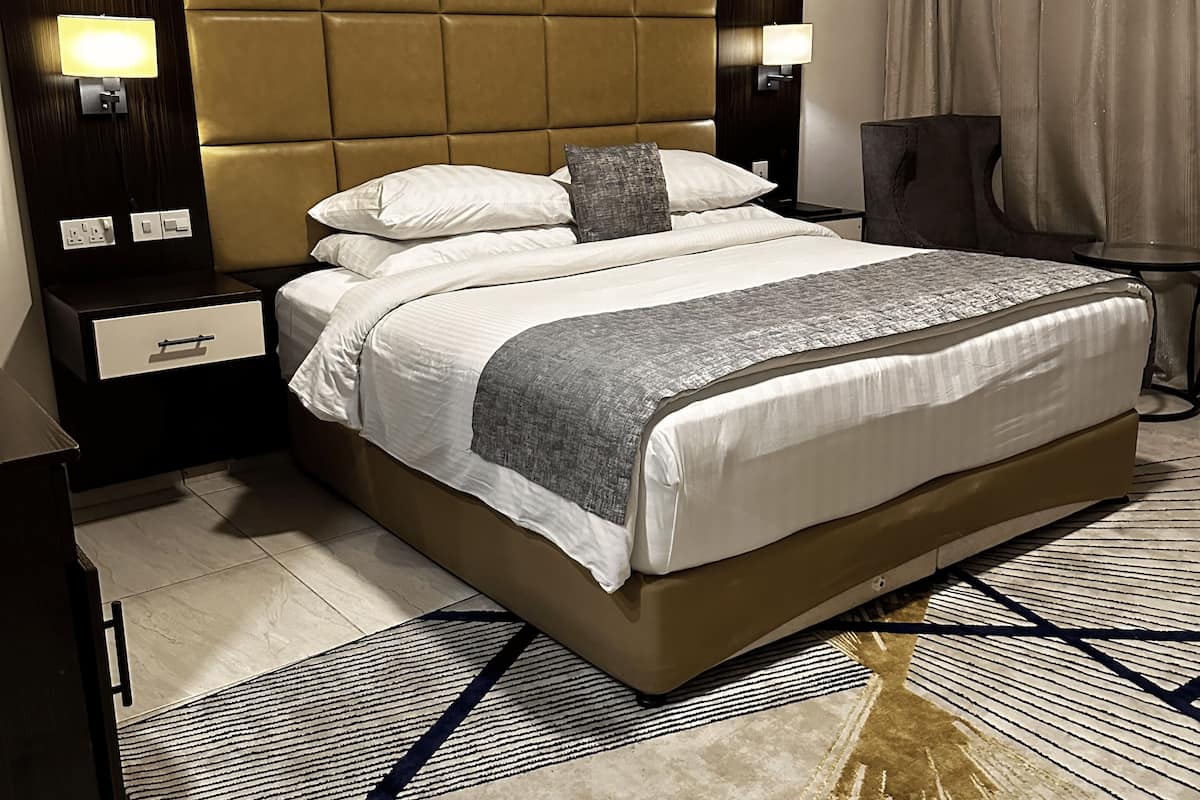 Una habitación de hotel moderna con una cama grande, un cabecero, dos mesitas de noche, un sillón y una alfombra estampada.