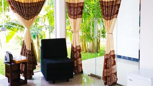 A hotel room with a bed, a chair, a small table with a kettle and teapots, and curtains with a pattern. - Rivonway Hotel Polonnaruwa (Giritale)