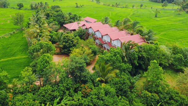 A large, multi-story building with a red roof, surrounded by lush green fields and palm trees. - Rivonway Hotel Polonnaruwa (Giritale)