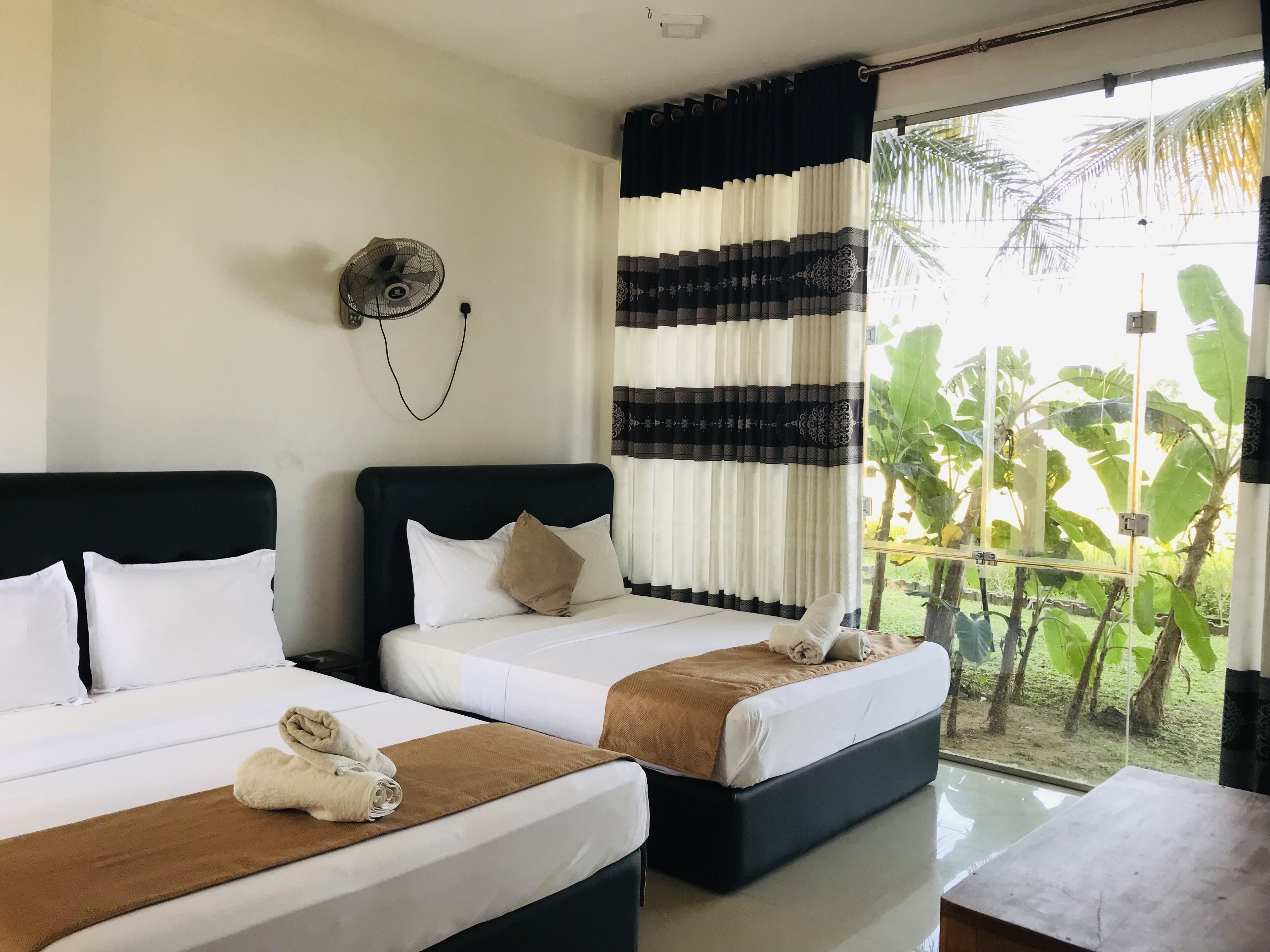 A hotel room with two beds, a ceiling fan, and a view of a garden with large leaves.