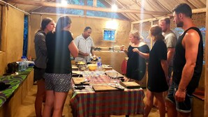 A group of people gathered around a table in a tented area with a rustic interior, featuring a checkered tablecloth, various food items, and a small window with a view. - Adannz view (Ella)